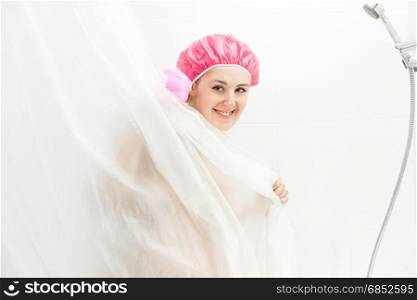 Portrait of cute smiling woman wearing shower cap having bath and hiding behind curtain