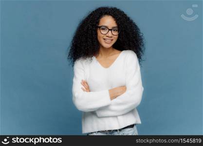Portrait of delighted pleasant looking curly female keeps arms folded on chest, has confident facial expression, charmin smile on face, wears white sweater and jeans, stands over blue background.