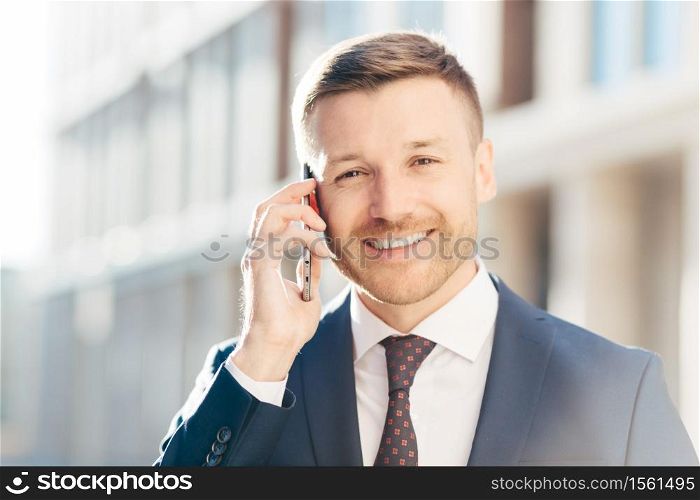 Portrait of delightful European businessman, wears elegant suit, talks on smart phone, deals with business partner, decide time of meeting, has positive expression. People, career, technology concept