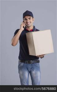 Portrait of delivery man carrying package while answering mobile phone against gray background