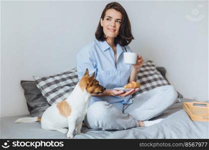 Portrait of dreamy brunette female sits crossed legs on comfortable bed in bedroom, looks thoughtfully aside, holds cup of coffee and croissant and her favourite jack russell terrier
