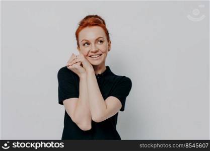 Portrait of dreamy happy ginger girl dressed casually looking aside at copy space with smile, adorable smiling red haired female holding hands under chin, standing isolated over grey studio background. Portrait of dreamy ginger girl looking aside at copy space with smile, holding hands under chin