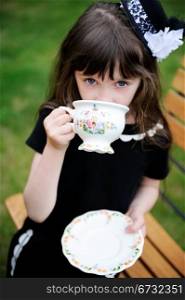 Portrait of elegant child girl in a black dress having a tea party outdoors, focus on a tea cup