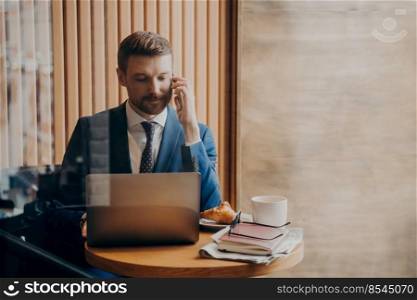 Portrait of elegant financier in stylish suit sitting in cafe with open laptop in front of him while talking on cellphone about work and business plan with concentrated look, verifying info on screen. Portrait of elegant finance man in stylish suit sitting in cafe with open laptop