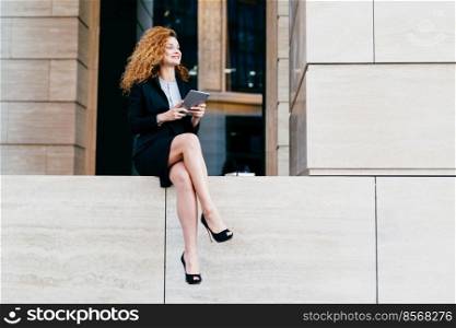 Portrait of elegant gorgeous woman in black suit and high-heeled shoes having slender long legs, sitting with modern tablet, looking aside with happy expression. Female office worker posing outdoor