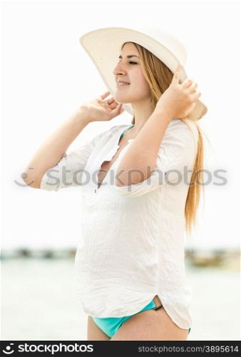 Portrait of elegant woman in hat standing on beach and looking at horizon
