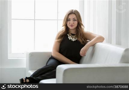 Portrait of elegant woman in long black dress relaxing on sofa next to big window