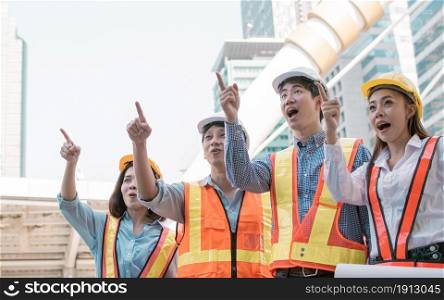 Portrait of engineering group wearing uniforms, safety helmets and point at something with surprising faces while standing outside near buildings. Construction Concept.