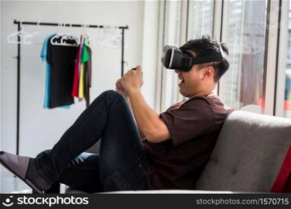 Portrait of excited Asian man on sofa play online game by VR glasses and wireless joystick in house. Happy guy play virtual gaming in condo. Leisure lifestyle concept.Stay home during covid19 pandemic