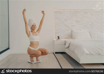 Portrait of excited young lovely caucasian woman squats on scales in bedroom at home, happy to achieved desired weight result, looking and feeling fit and healthy. Weight loss and dieting concept. Portrait of excited young lovely caucasian woman squats on scales in bedroom
