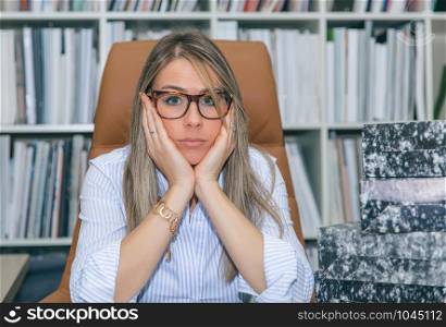 Portrait of exhausted blonde secretary looking at camera with a lot of work waiting in her desktop. Exhausted secretary with lot of work in office