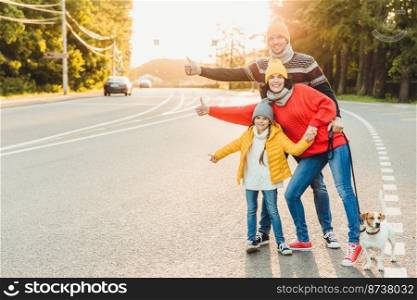 Portrait of family: mother, father and little daughter walk with pet, stand on road, raise thumbs as show ok sign,express approval or agreement, have good mood after spending time on fresh air