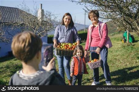 Portrait of family with fresh organic apples in a wicker basket posing while cute boy taking a photo with electronic tablet. Three different generations concept.. Family with apples in basket posing to photo