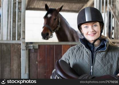Portrait Of Female Owner Holding Saddle In Stable With Horse