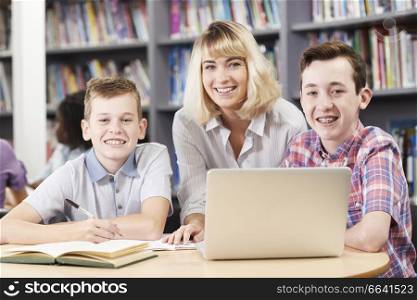 Portrait Of Female Teacher Helping Two Male High School Students Working At Laptop In Library