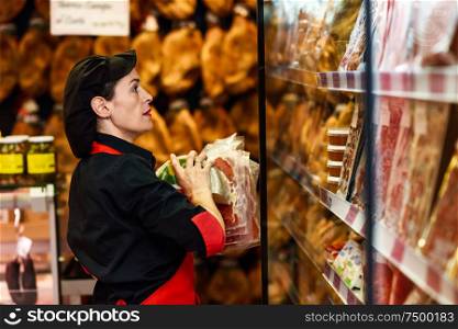 Portrait of female worker taking products in butcher shop. Refrigerated display case for sausage packages, ham and cheese. Portrait of female worker taking products in butcher shop