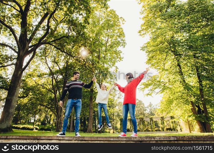 Portrait of friendly affectionate family have fun together, spend free time outdoors, stand against green trees in park, try to rock small child in hands. Three family memebers play on nature