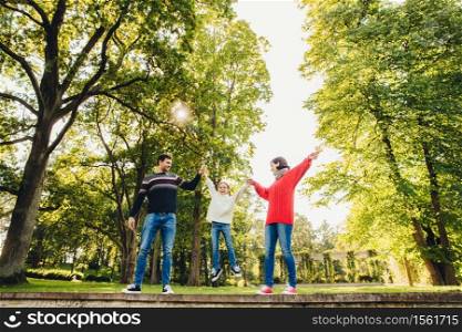 Portrait of friendly affectionate family have fun together, spend free time outdoors, stand against green trees in park, try to rock small child in hands. Three family memebers play on nature
