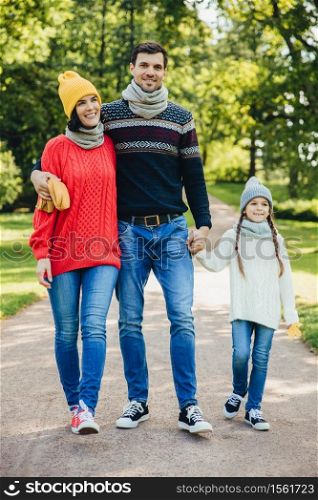 Portrait of friendly family walk across park. Beautiful brunette female in knitted yellow hat and sweater, handsome man embraces his wife and holds daughter`s hand, enjoy autumn weather and fresh air