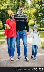 Portrait of friendly family walk across park. Beautiful brunette female in knitted yellow hat and sweater, handsome man embraces his wife and holds daughter`s hand, enjoy autumn weather and fresh air