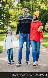 Portrait of friendly family walk across park. Beautiful brunette female in knitted yellow hat and sweater, handsome man embraces his wife and holds daughter s hand, enjoy autumn weather and fresh air