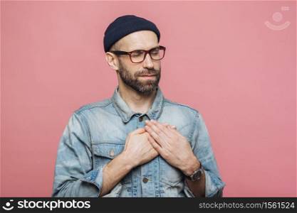 Portrait of friendly looking bearded male with closed eyes, keeps hands on chest, wears denim jacket, glasses and hat, isolated over pink background. Middle aged unshaven man gestures indoor
