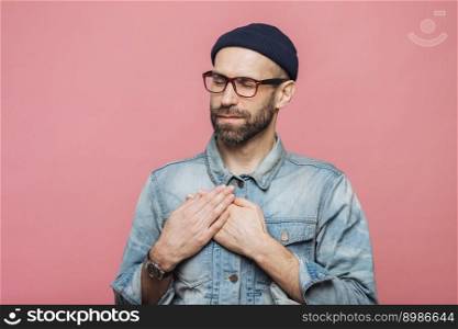 Portrait of friendly looking bearded male with closed eyes, keeps hands on chest, wears denim jacket, glasses and hat, isolated over pink background. Middle aged unshaven man gestures indoor