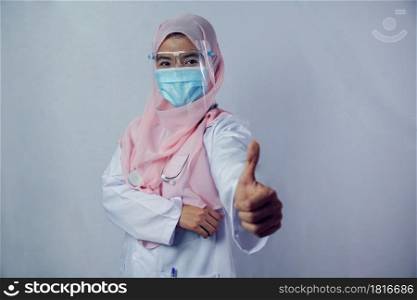 Portrait of friendly, smiling confident Muslim woman doctor in hijab dress wear mask and face shield holding a stethoscope and thumb up looking at camera on white background