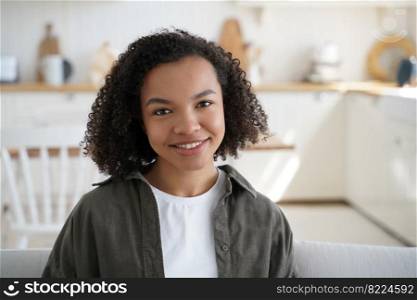 Portrait of friendly smiling young pretty mixed race girl with afro hairstyle looking at camera at home, modern happy teen lady sits on sofa, enjoying lazy weekend in cozy homey house on a sunny day.. Friendly smiling young pretty mixed race girl with afro hairstyle looking at camera in cozy home