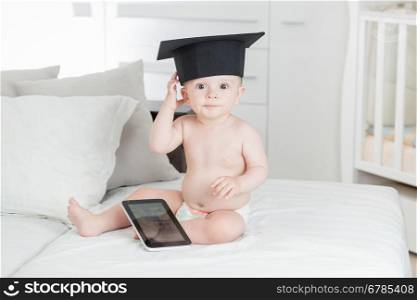 Portrait of funny baby boy in graduation cap using digital tablet