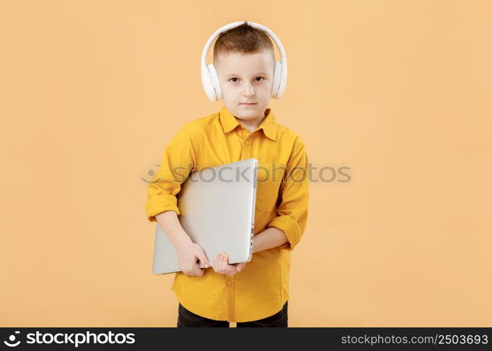 Portrait of funny clever school boy with headphones and laptop in yellow shirt. Yellow studio background. Education. Looking at camera. High quality photo.. Portrait of funny clever school boy with headphones and laptop in yellow shirt. Yellow studio background. Education. Looking at camera. High quality photo