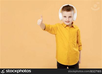 Portrait of funny clever school boy with headphones in yellow shirt. Yellow studio background. Education. Looking, smiling and shows a thumb up at camera. High quality photo.. Portrait of funny clever school boy with headphones in yellow shirt. Yellow studio background. Education. Looking, smiling and shows a thumb up at camera. High quality photo