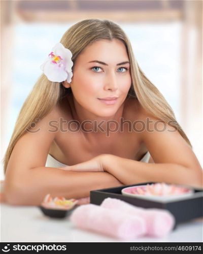 Portrait of gentle blond female with orchid flower in hair lying down on massage table, enjoying beauty treatment in luxury spa salon