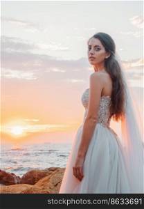 Portrait of girl in wedding luxury dress watching sunrise on sea shore. Bride on a rocks. Beautiful waves near to her. Woman enjoying happy moments with nature.