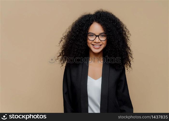 Portrait of glad curly haired young businesswoman with toothy smile, has delighted expression, wears spectacles and formal clothes, ready for meeting, poses over brown background. Lady in black