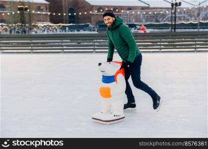 Portrait of glad handsome middle aged male skates on ice rink with skate aid, going to show good results, has pleasant smile, happy to spend spare time on fresh winter air during nice weather