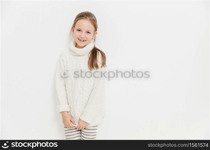 Portrait of glad satisfied girl wears warm white knitted sweater, has joyful expression, stands against studio background with blank copy space. Adorable female child enjoys leisure time. Monochrome
