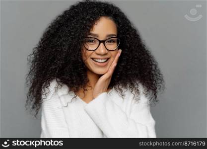 Portrait of good looking Afro woman touches cheek, smiles happily, enjoys receiving compliment, feels shy, dressed in white casual sweater, poses indoor. People, positive emotions and feelings concept