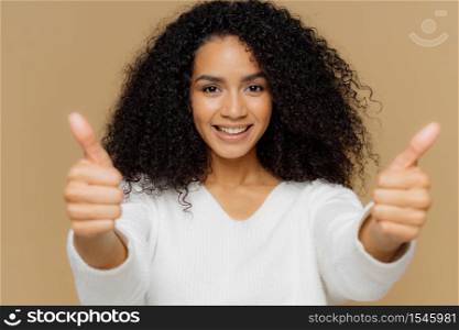 Portrait of good looking female raises thumbs, shows like gesture in camera, enjoys nice presentation, dressed in casual clothes, poses against brown background. Approval and recommendation concept