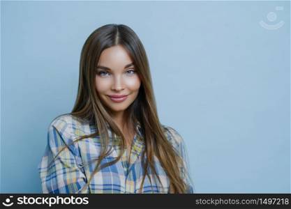 Portrait of good looking female student poses against blue studio wall, looks at camera with charming smile, dresses checkered shirt, poses for university photo, feels optimistic, wears make up