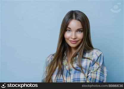 Portrait of good looking female student poses against blue studio wall, looks at camera with charming smile, dresses checkered shirt, poses for university photo, feels optimistic, wears make up