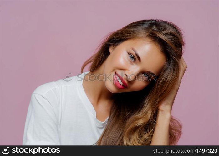 Portrait of good looking pleased female model tilts head, has toothy smile shows white perfect teeth, wears casual jumper, touches hair, shows her beauty, being in good mood, isolated on purple wall