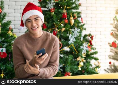 Portrait of good-looking smart Teenager young asian adult boy wearing brown sweater using his mobile phone for social media while waiting her girlfriens for Christmas celebration in living room with christmas decoration.