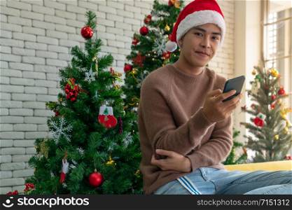 Portrait of good-looking smart Teenager young asian adult boy wearing brown sweater using his mobile phone for social media while waiting her girlfriens for Christmas celebration in living room with christmas decoration.
