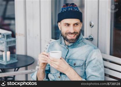 Portrait of good looking smiling bearded male, wears warm hat and denim jacket, drinks aromatic tea, has happy expression, sits against terrace interior. People, leisure and recreation concept