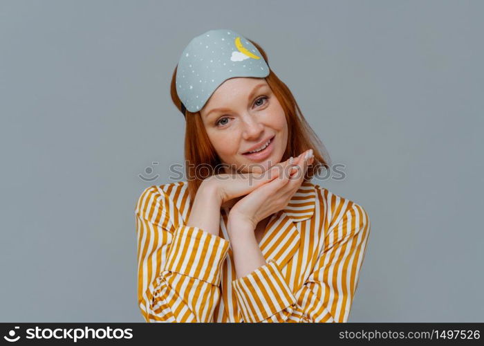 Portrait of good looking woman gets enough amount of sleep, enjoys pleasant relaxation, tilts head and keeps hands under chin, wears blindfold and striped pajamas, isolated over grey background