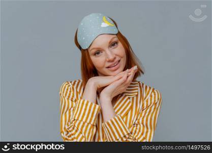 Portrait of good looking woman gets enough amount of sleep, enjoys pleasant relaxation, tilts head and keeps hands under chin, wears blindfold and striped pajamas, isolated over grey background