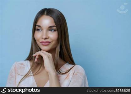 Portrait of good looking woman touches chin and has thoughtful expression, looks away, isolated over blue background, blank space for your advertisement or promotion. Romance and beauty concept