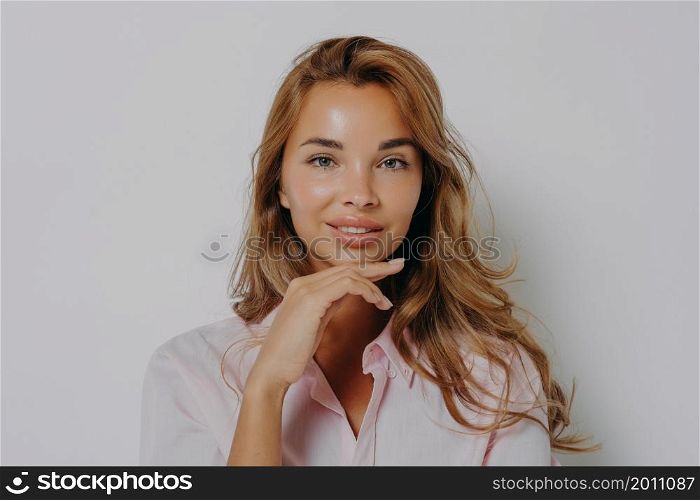 Portrait of good looking woman with long hair healthy skin minimal makeup touches chin gently looks directly at camera wears casual shirt isolated over grey background. Women and beauty concept. Portrait of good looking woman with long hair healthy skin minimal makeup touches chin gently