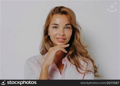 Portrait of good looking woman with long hair healthy skin minimal makeup touches chin gently looks directly at camera wears casual shirt isolated over grey background. Women and beauty concept. Portrait of good looking woman with long hair healthy skin minimal makeup touches chin gently